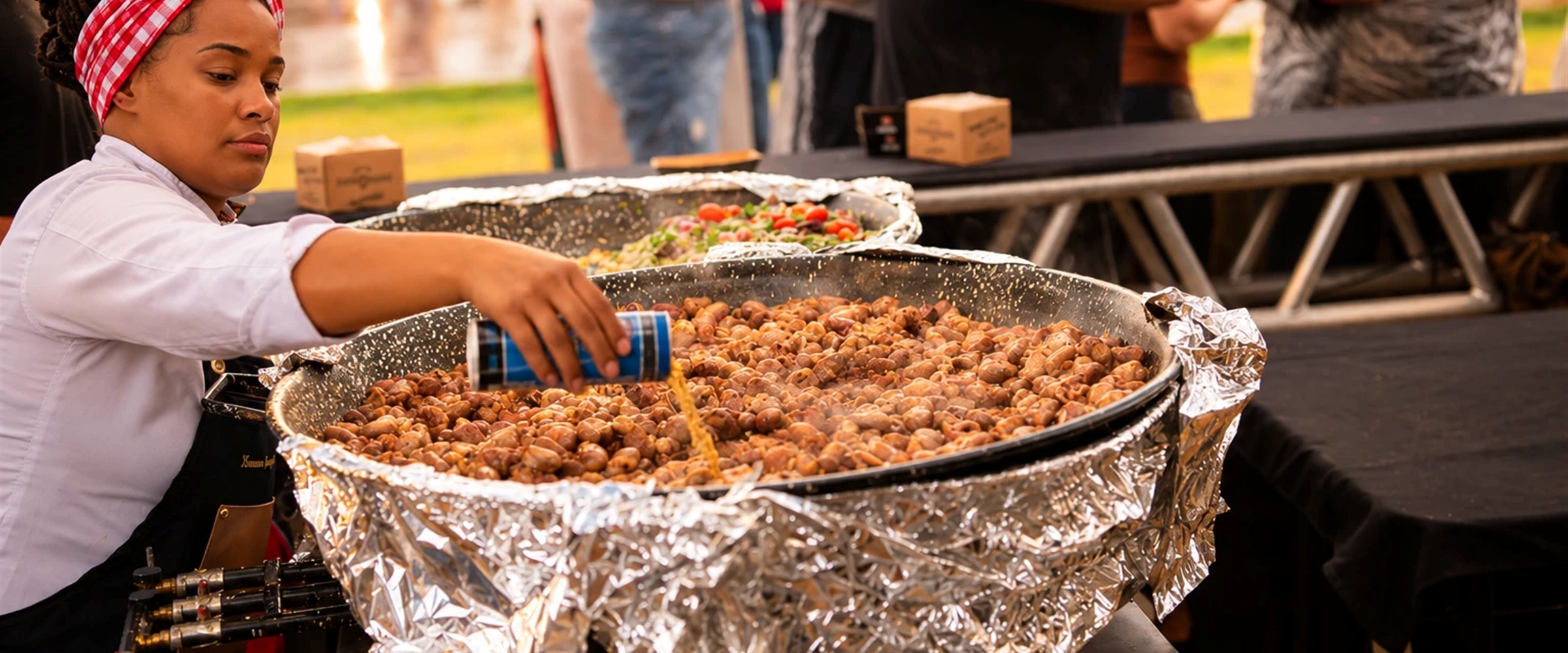 Cozinheira temperando uma grande panela coberta com papel alumínio em um evento ao ar livre
